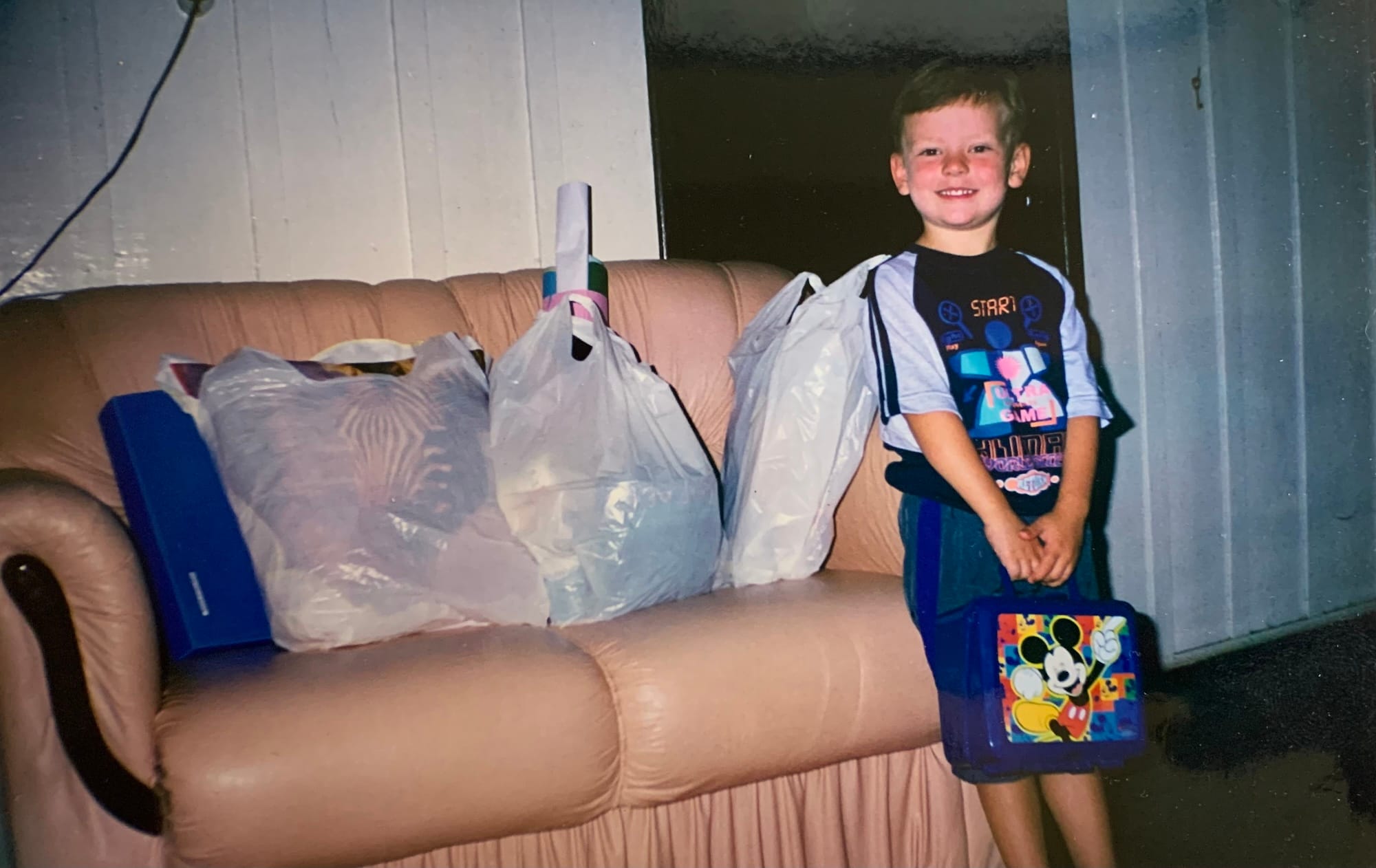 A white kid smiles while holding a blue lunchbox next to a beige sofa full of school supplies.