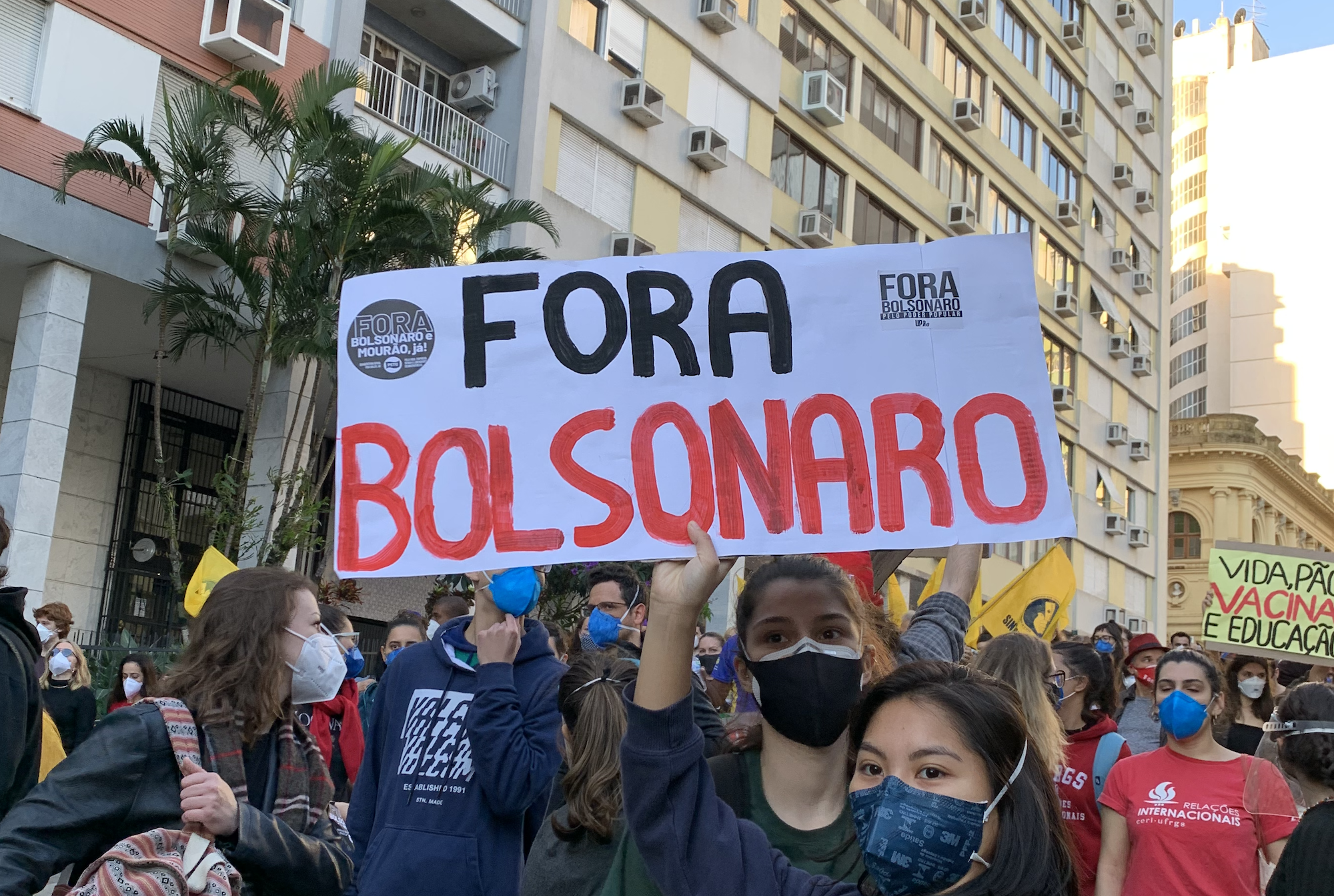 A young lady holds a sign that reads "Down with Bolsonaro" in Portuguese. She's surrounded by other young people in a town square on a sunny day.